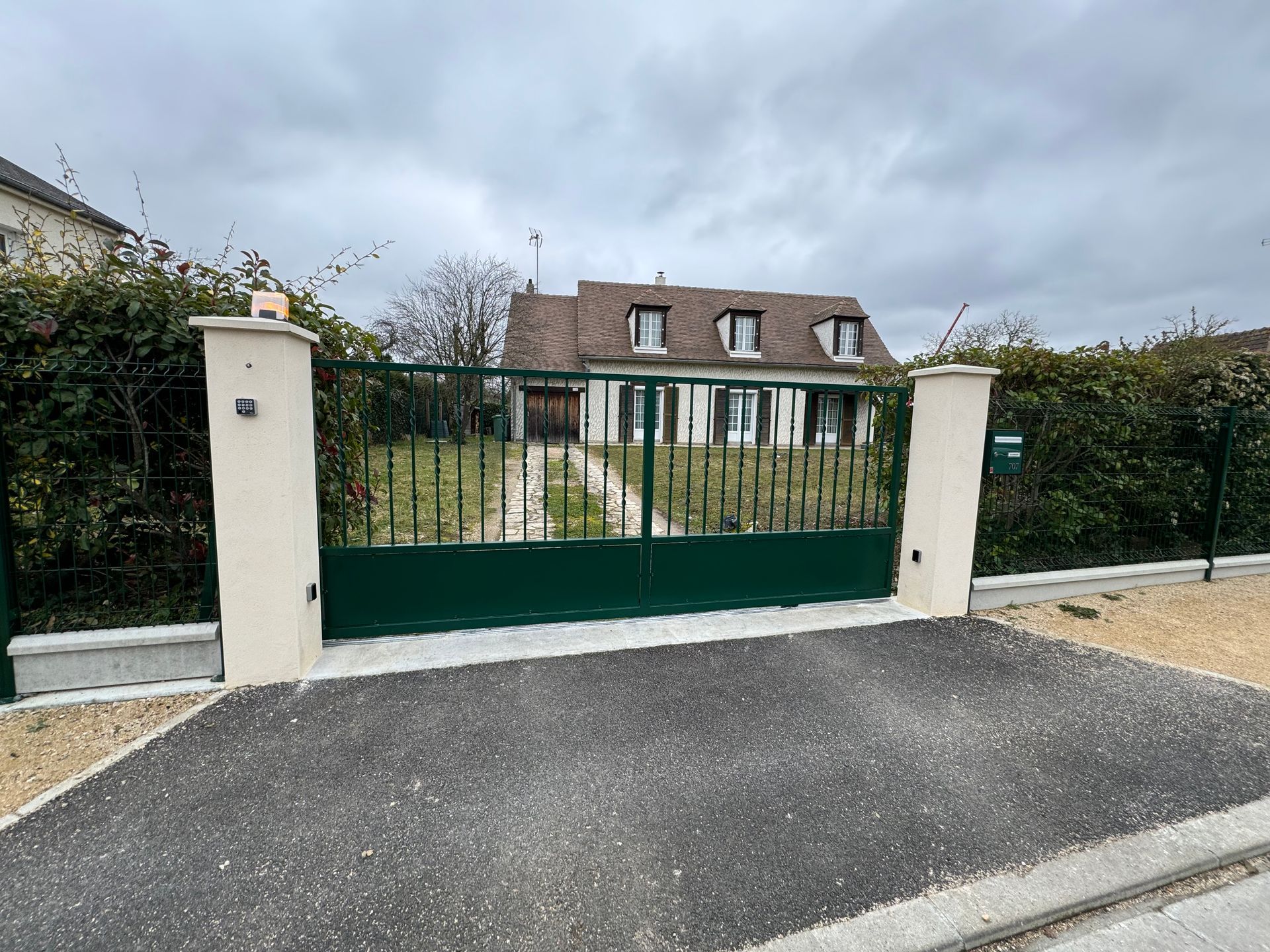 Un portail vert mène à une maison avec une allée de gravier et des haies environnantes, par temps couvert.