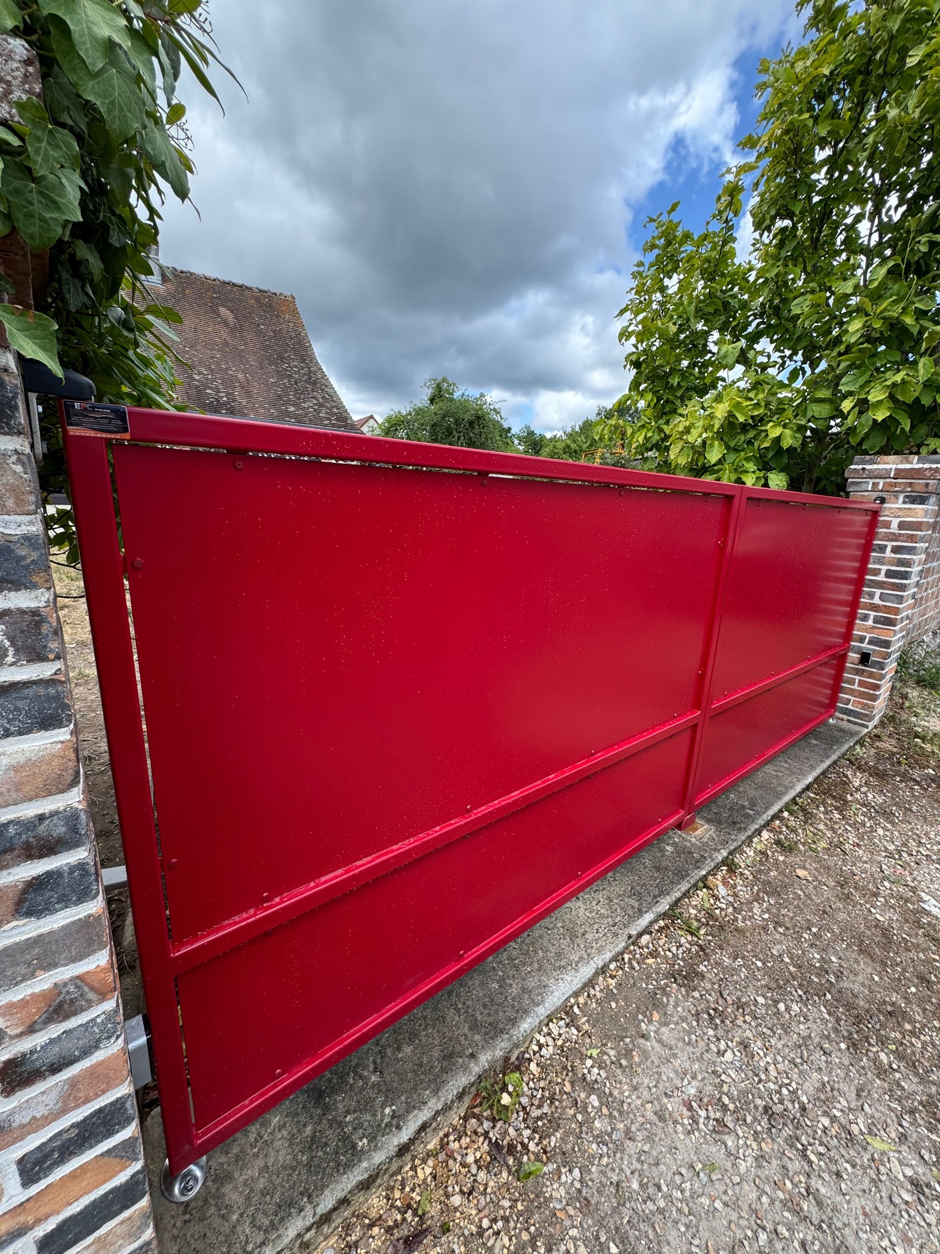 Portail en métal rouge encastré dans des murs de briques, ciel nuageux au-dessus.
