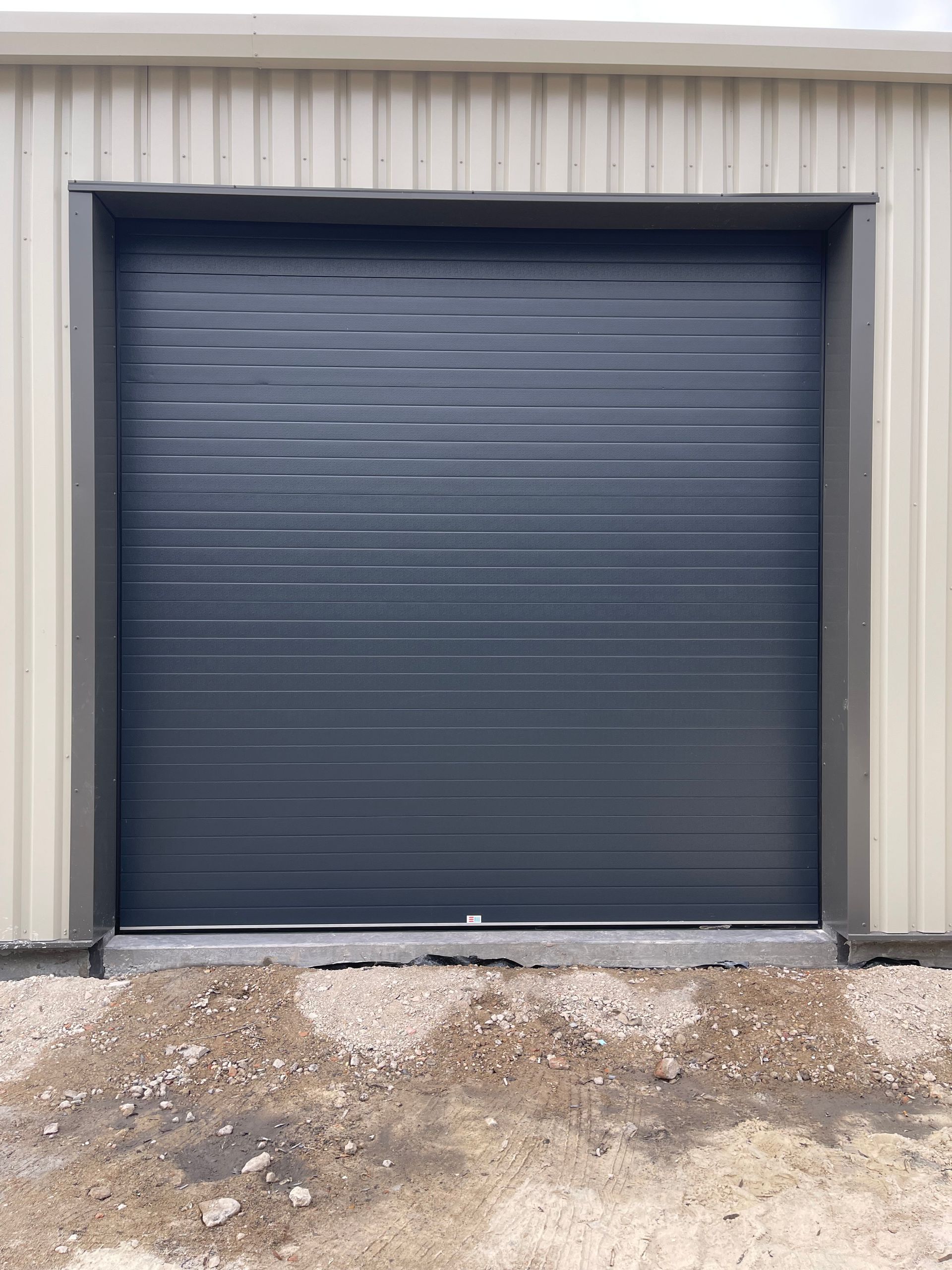 Porte de garage à panneaux horizontaux gris foncé dans un bâtiment métallique beige, avec une base en béton.
