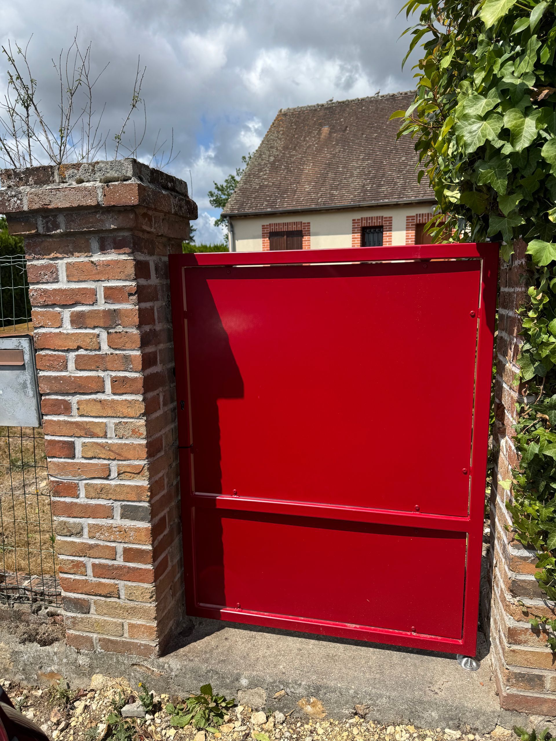 Portail en métal rouge enchâssé dans des piliers de brique, devant une maison.
