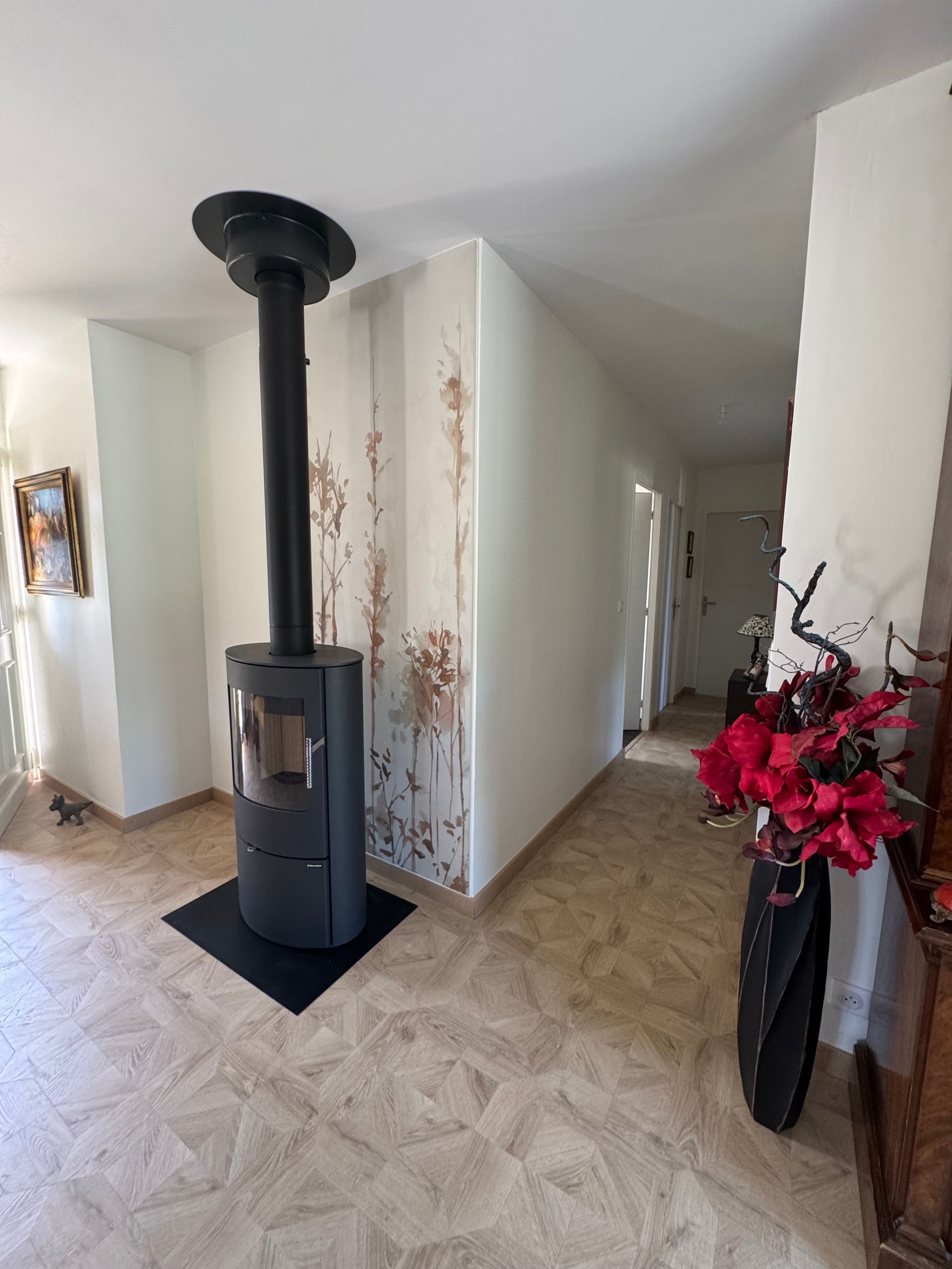 Dans une pièce aux murs et au sol clairs, un poêle à bois noir moderne est installé ; un couloir se trouve à droite.