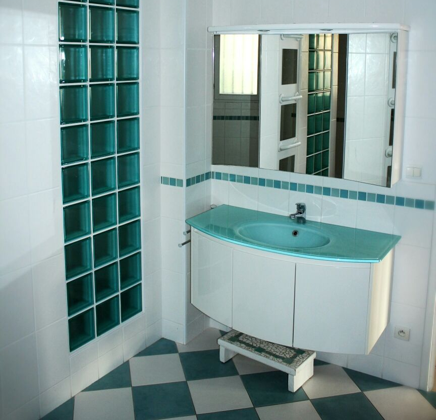 Salle de bains avec carrelage turquoise et blanc, accents de pavés de verre et lavabo incurvé.