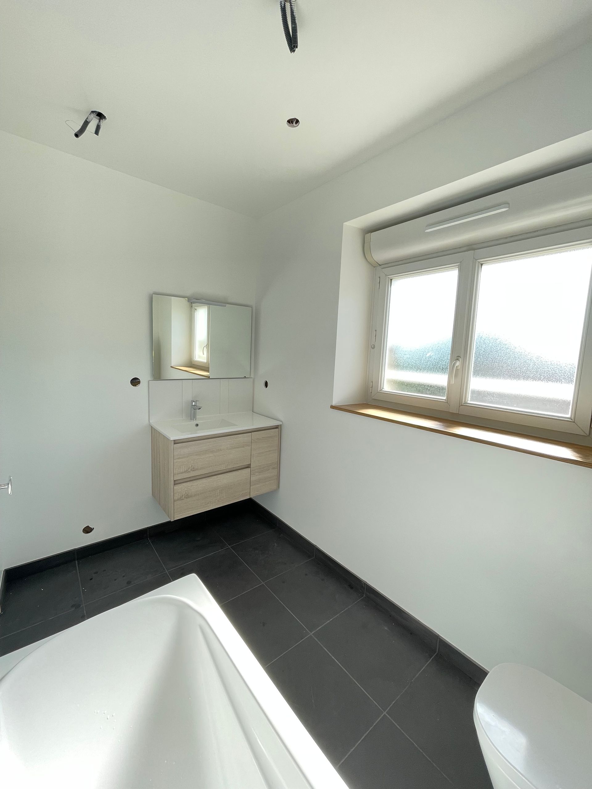 Salle de bains avec baignoire blanche, meuble-lavabo, fenêtre et carrelage foncé au sol.