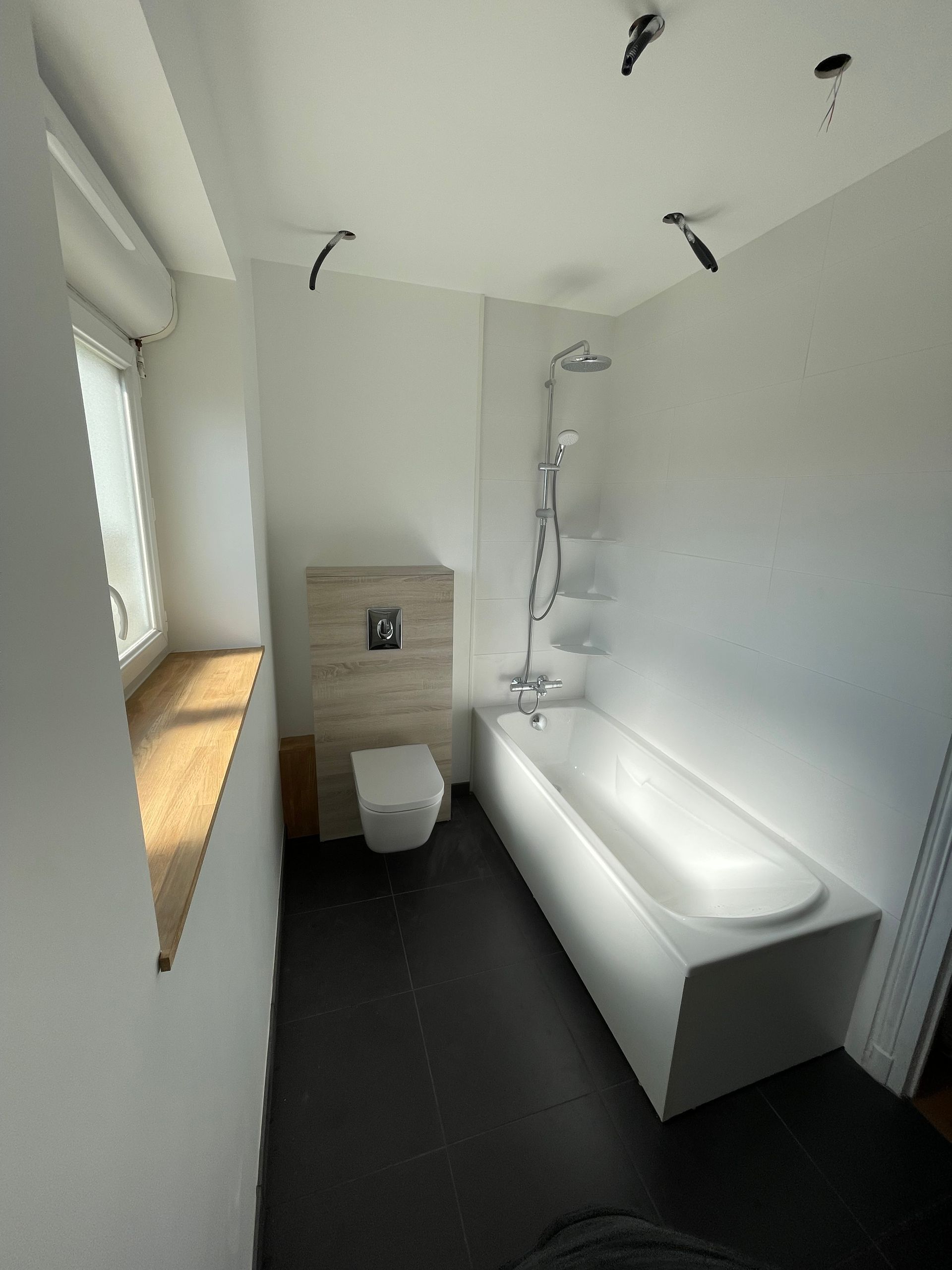 Salle de bains avec murs blancs, sol noir, toilettes, baignoire, pommeau de douche, fenêtre.