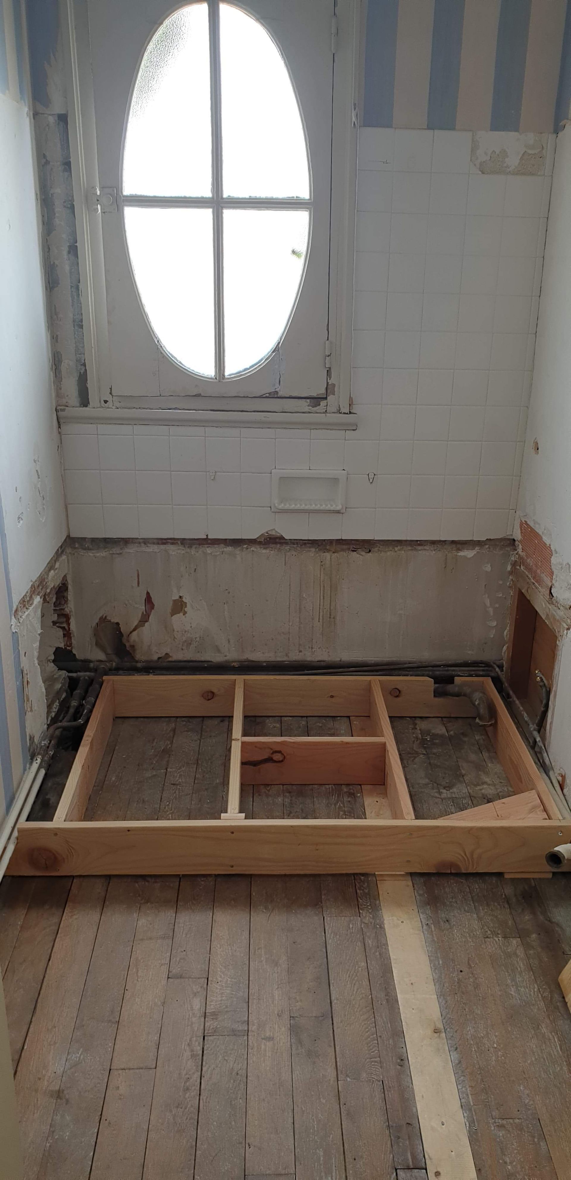 Intérieur rénové : fenêtre ovale, parquet, ossature bois partiellement construite au sol.