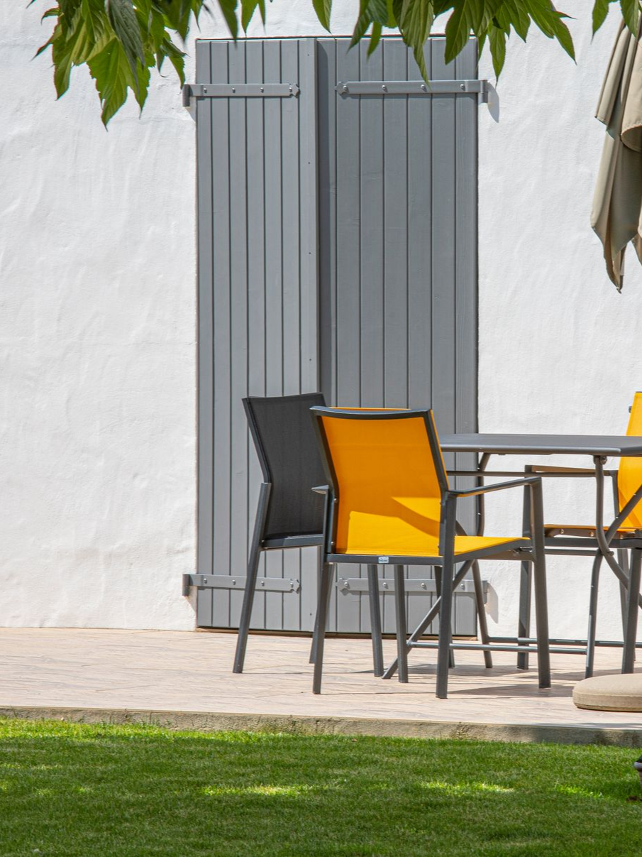 Espace salon extérieur avec volets gris, deux chaises à dossiers orange et une pelouse verte.