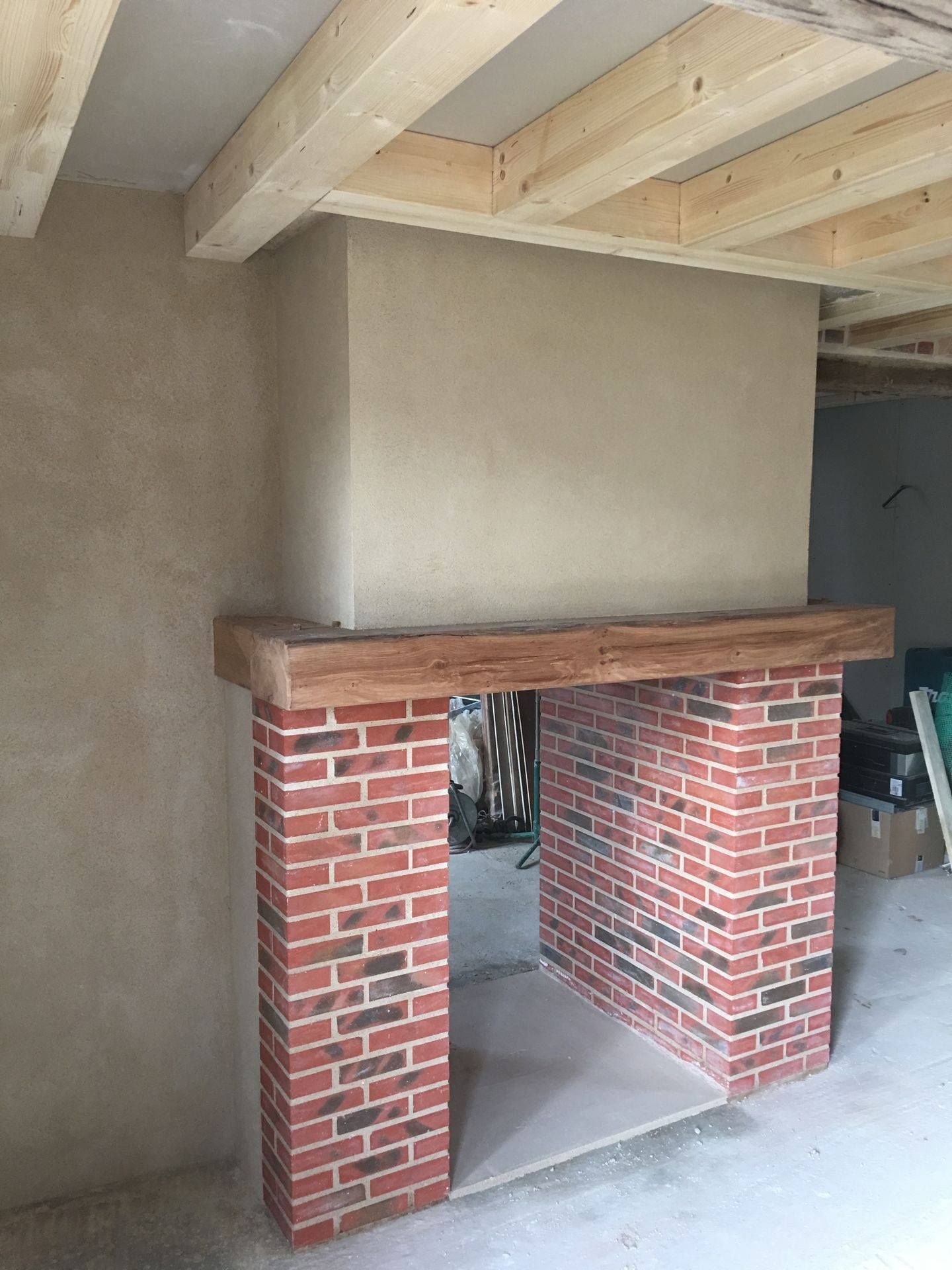 Cheminée en briques avec manteau en bois, installée dans une pièce aux poutres apparentes au plafond et aux murs plâtrés.