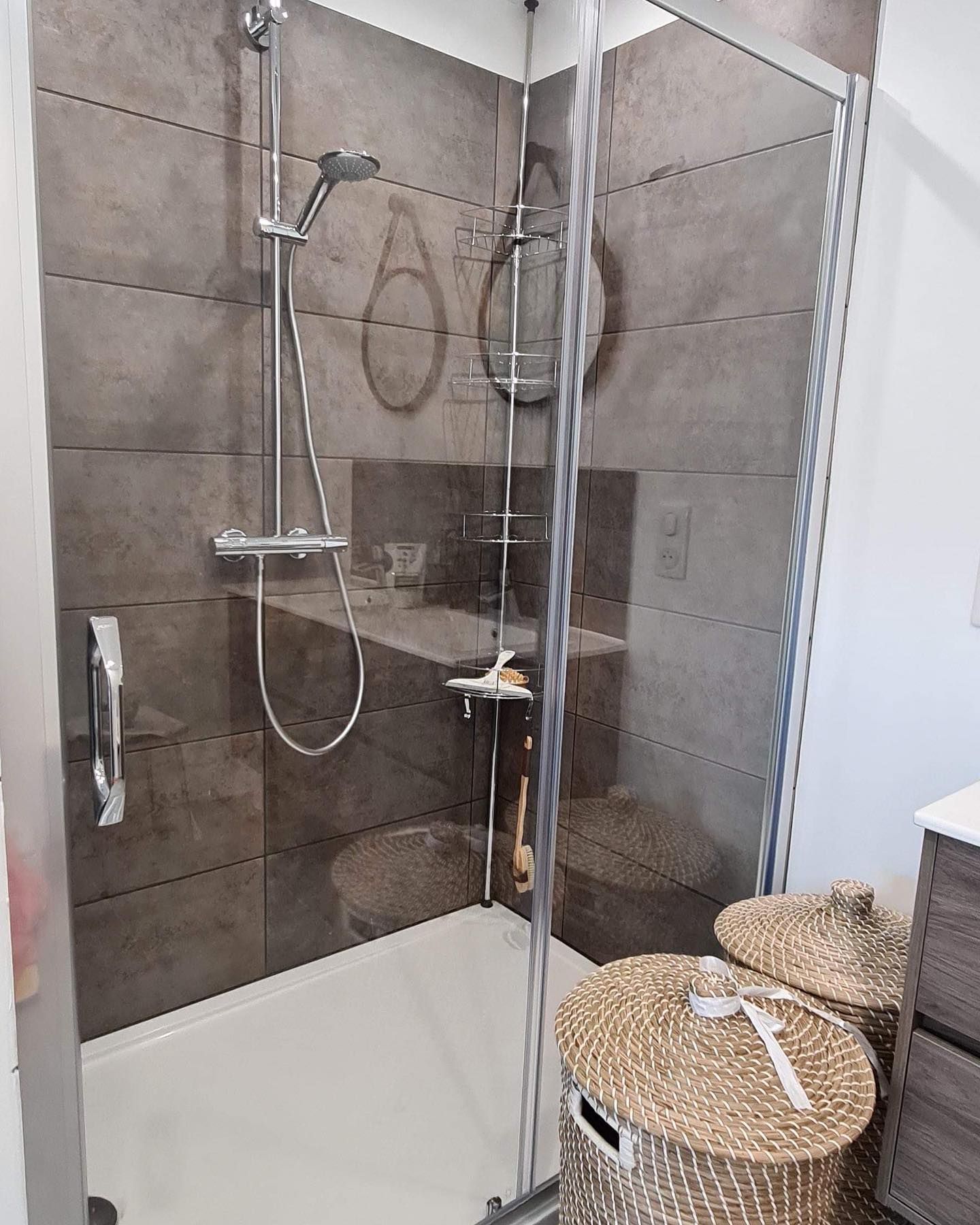 Cabine de douche avec carrelage gris, porte en verre et robinetterie chromée. Deux paniers en osier sont disposés à l'extérieur.