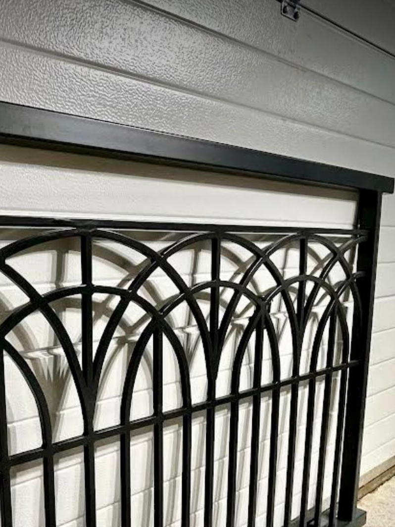 Clôture en fer forgé noir avec sommet arqué et barreaux verticaux, montée sur un mur blanc.