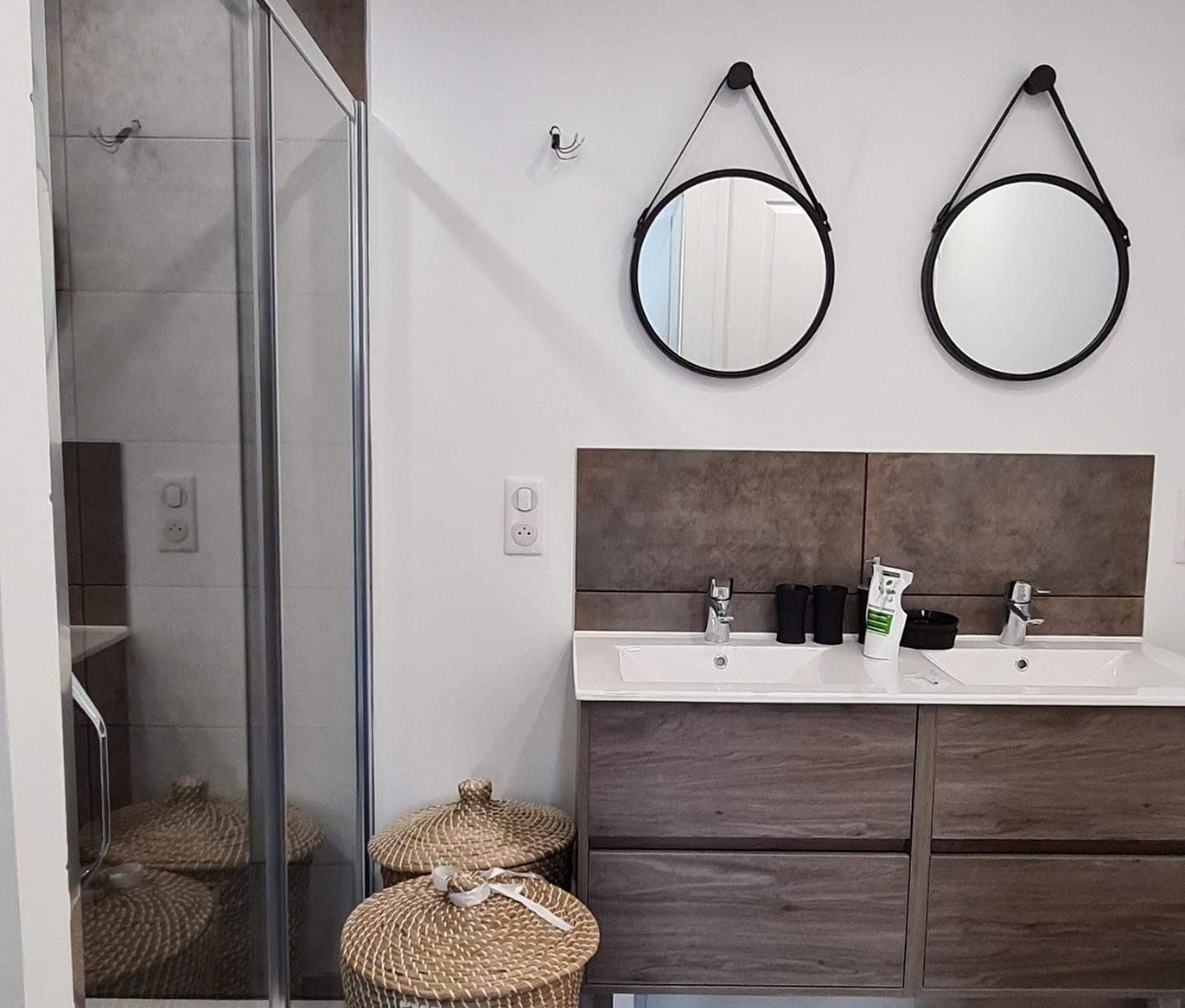 Salle de bains avec douche, double vasque, miroirs ronds et paniers tressés.