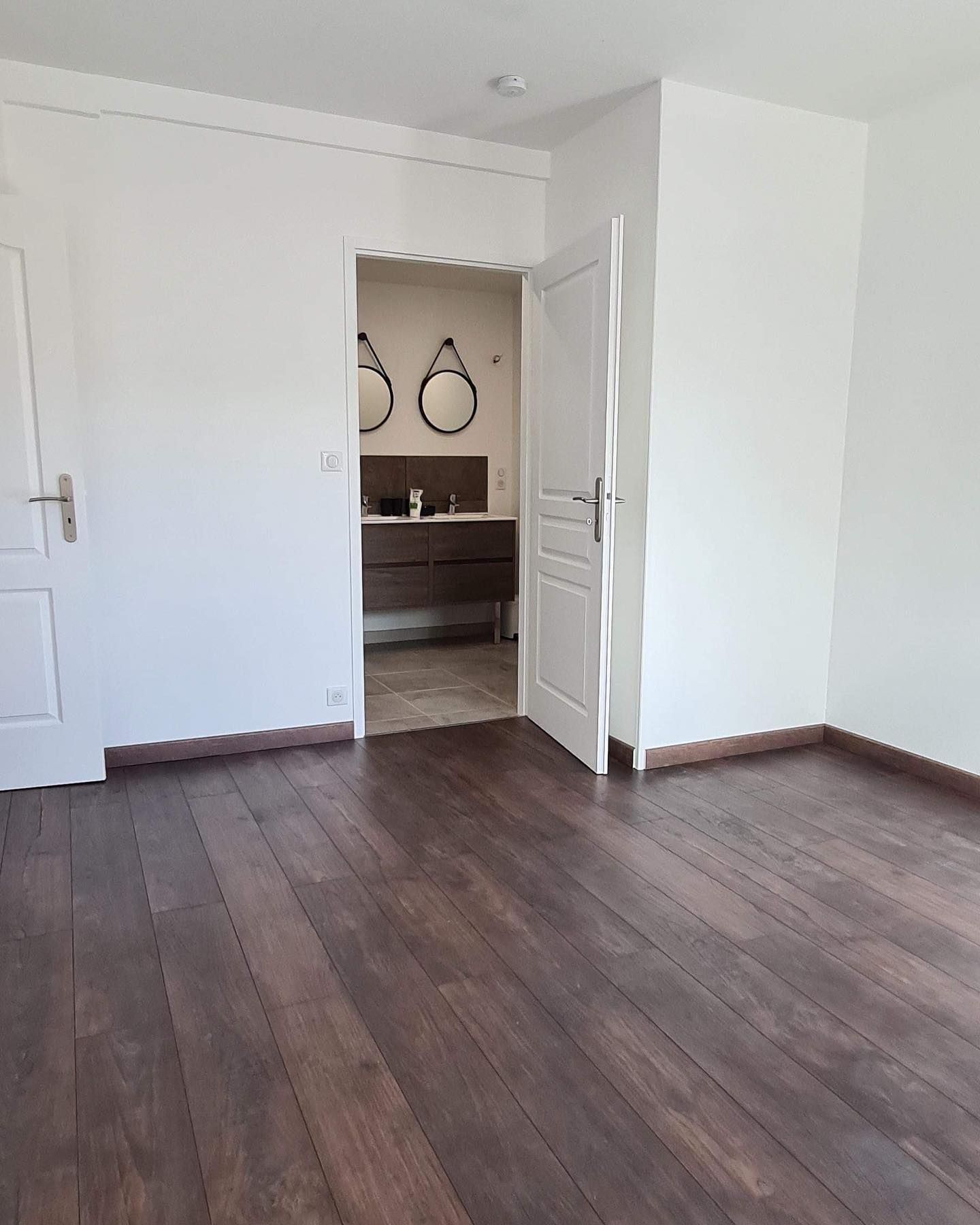Chambre vide avec parquet, murs et portes blancs, porte donnant sur la salle de bains avec miroirs.