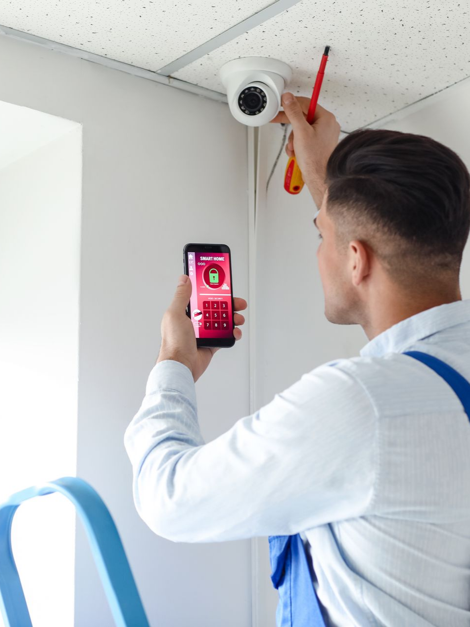 Un homme installe une caméra de sécurité, tient un téléphone affichant une application de sécurité, travaille à l'intérieur.