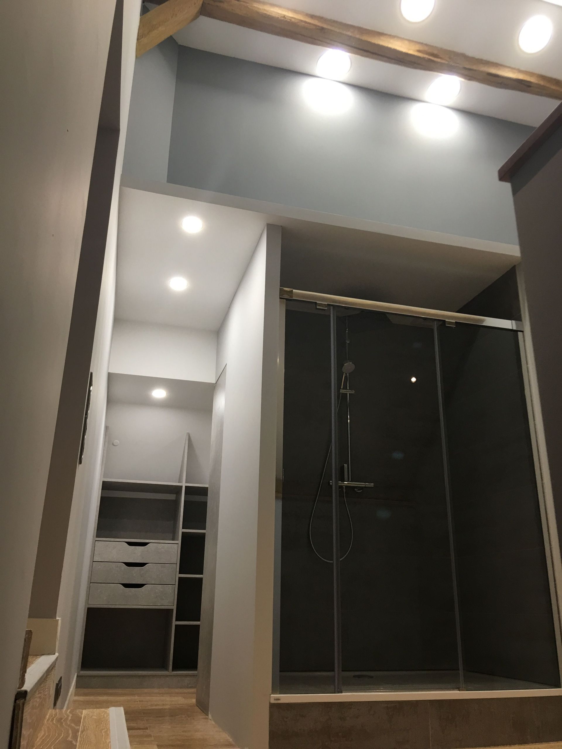Couloir étroit avec dressing et douche vitrée. Murs gris, éclairage encastré et poutres apparentes.