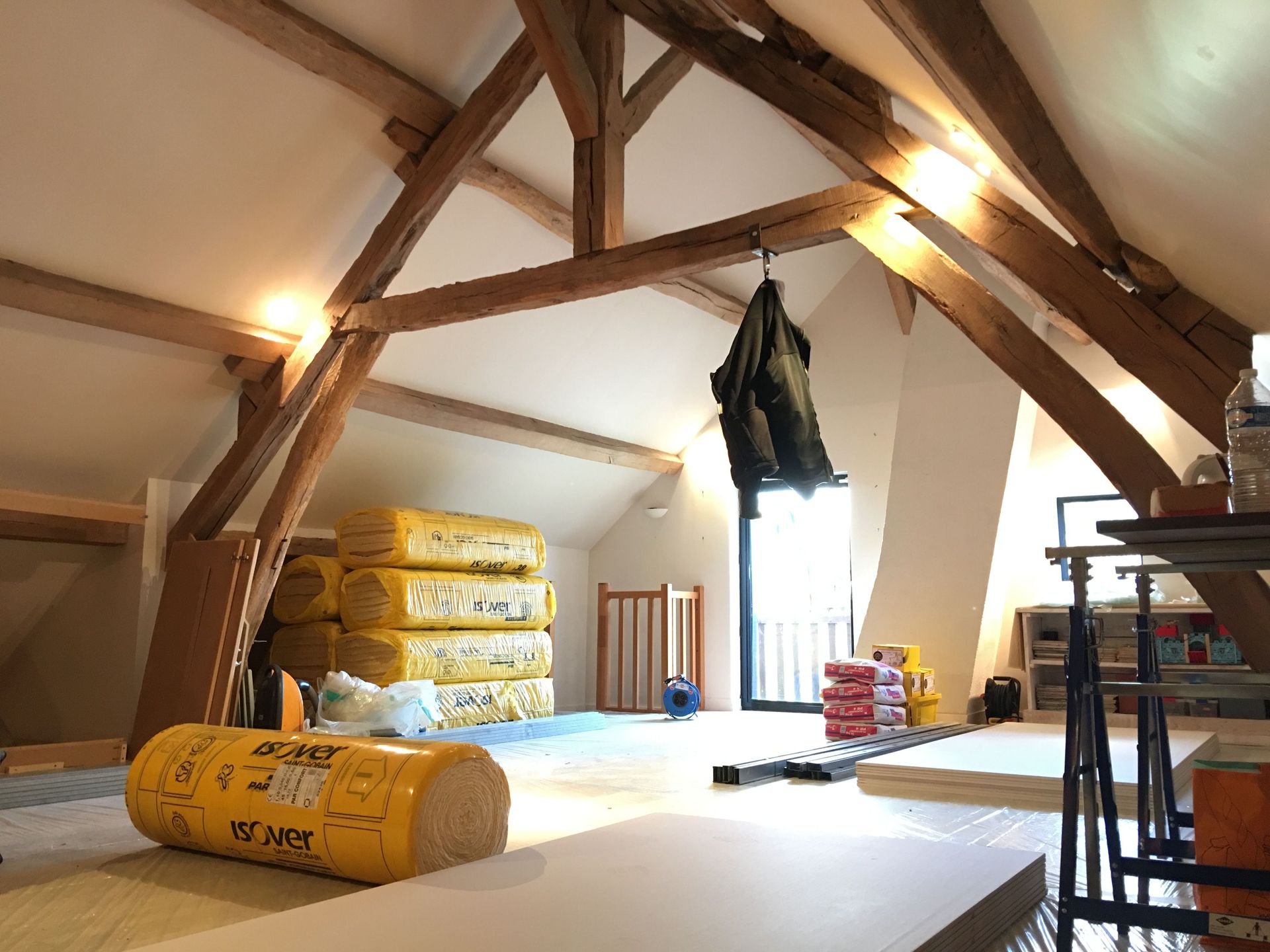 Grenier en cours d'aménagement avec poutres apparentes, rouleaux d'isolant et matériaux de construction.