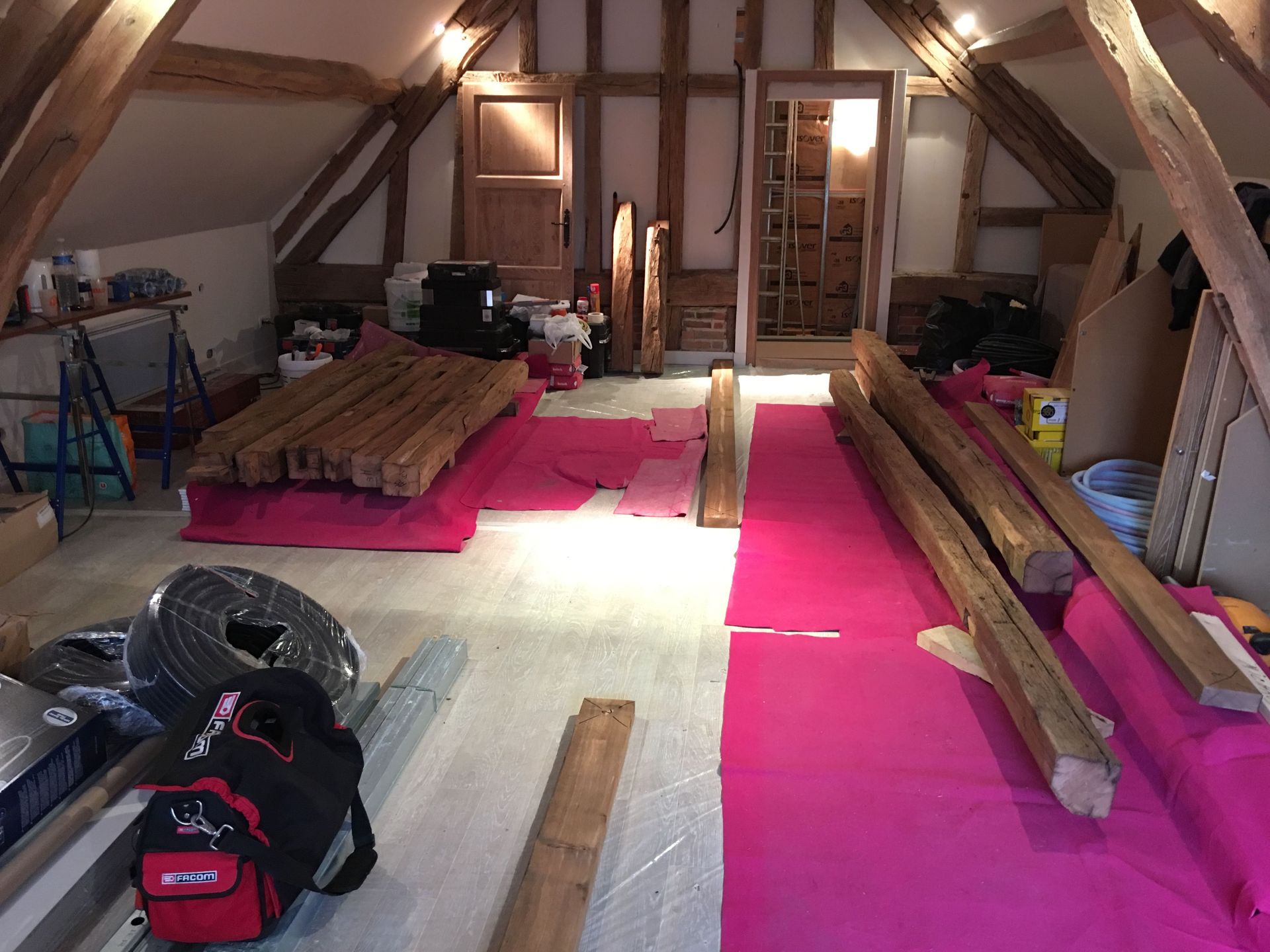 Grenier en travaux, avec poutres apparentes, bâches de protection roses et matériaux de construction.