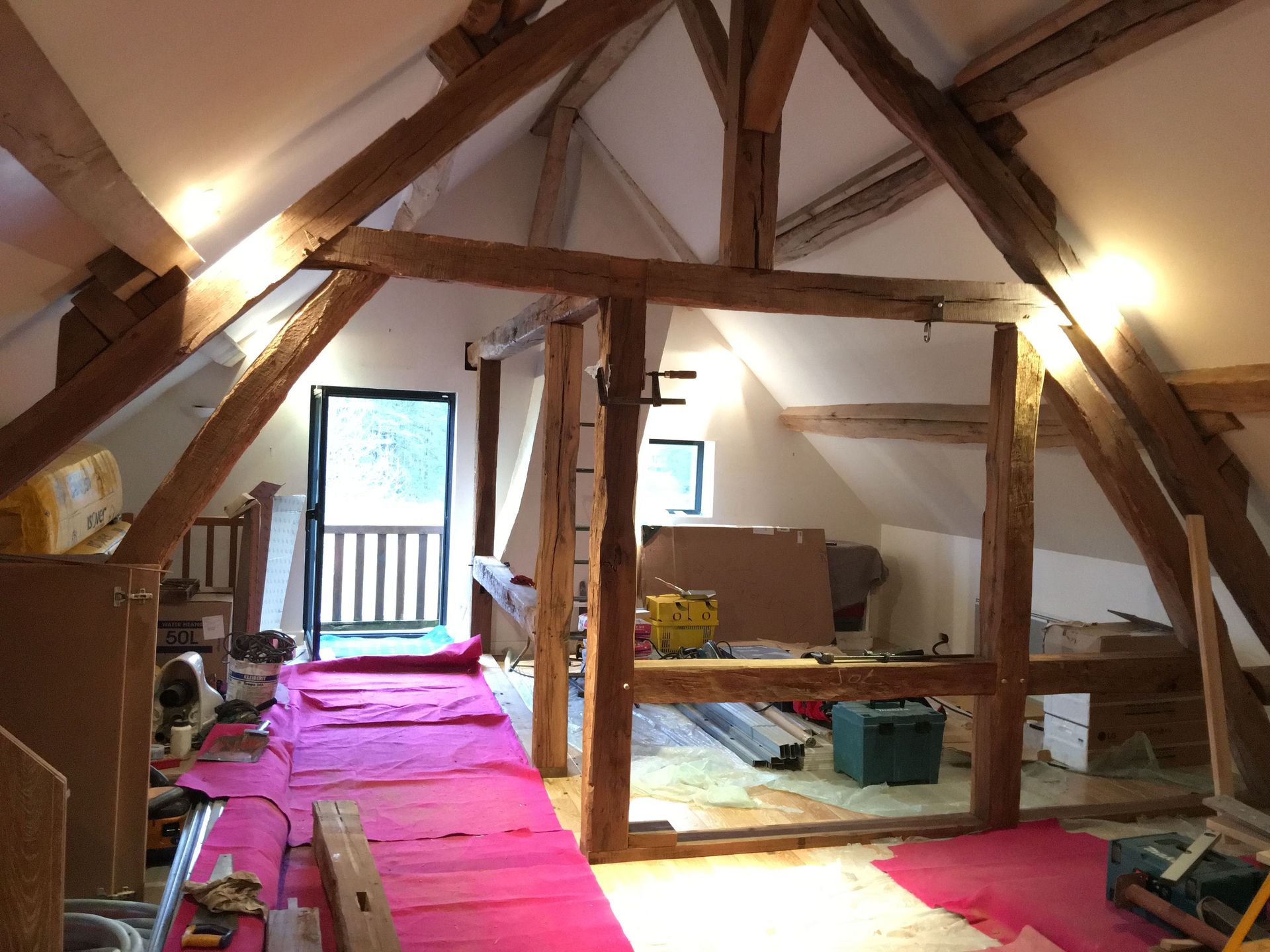 Grenier rénové avec poutres apparentes, revêtement de sol rose, porte ouverte et outils de construction.
