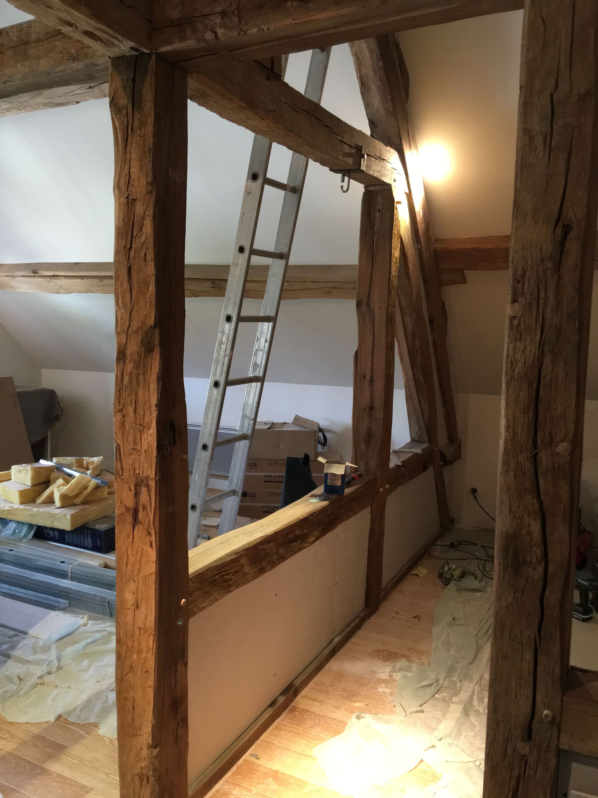 Vue intérieure d'une pièce en construction avec des poutres en bois apparentes, une échelle et des murs blancs.