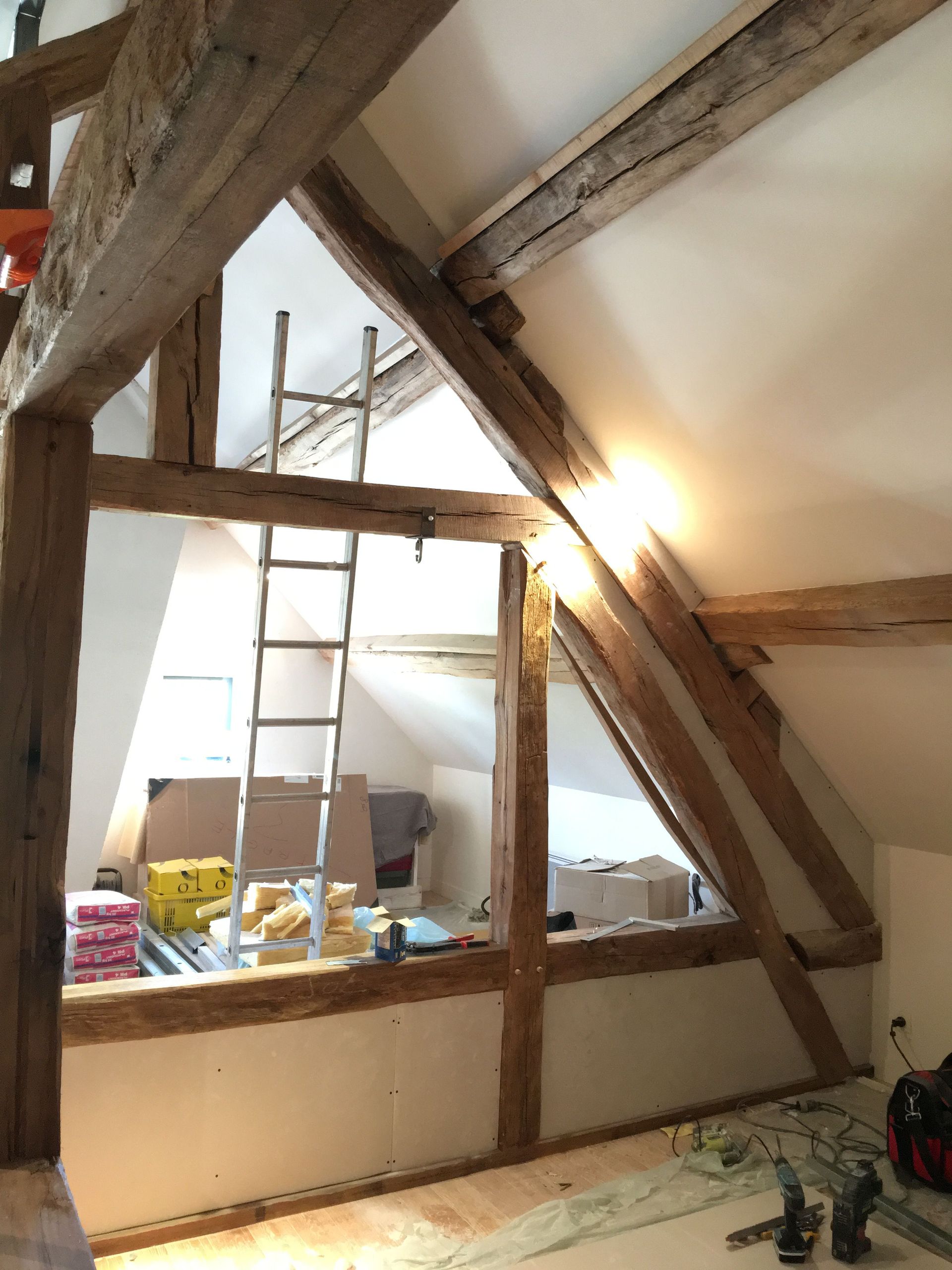 Vue intérieure d'une pièce en construction avec poutres en bois apparentes, cloisons sèches et une échelle.