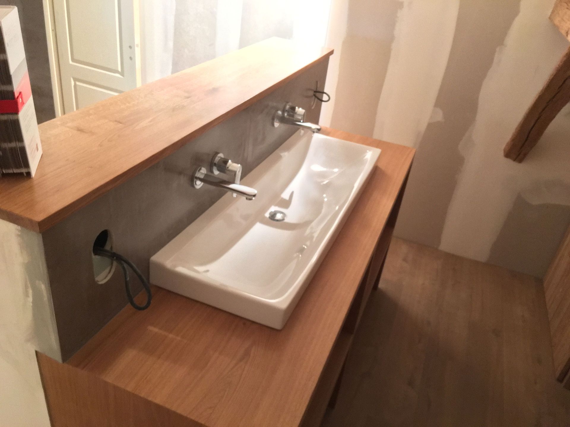 Meuble-lavabo en bois avec deux robinets et un lavabo rectangulaire blanc. Mur gris.
