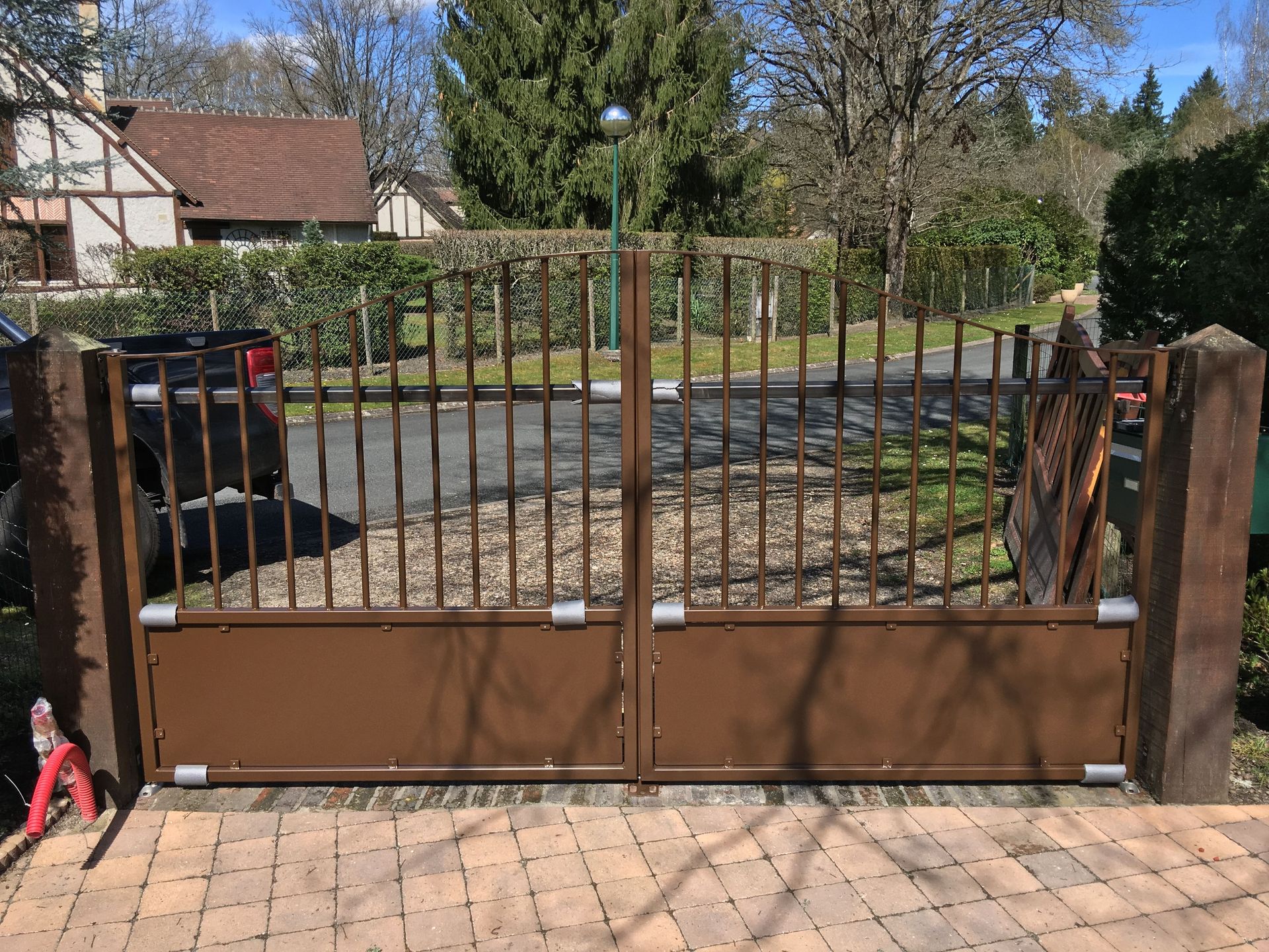 Portail en métal marron devant une maison, posé sur des pavés de brique, avec des barreaux verticaux et un éclairage central.