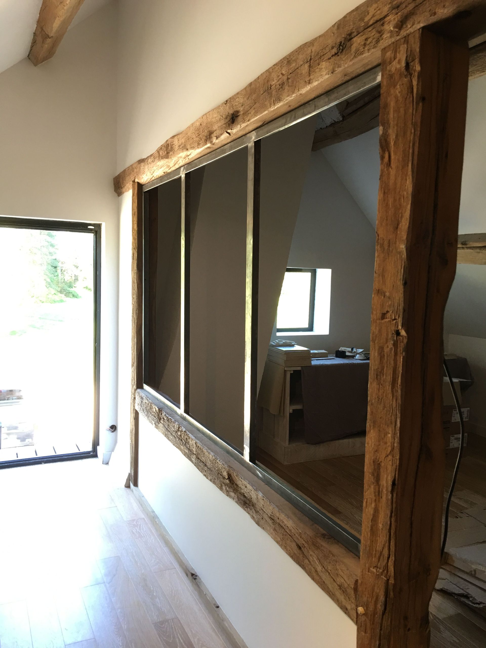 Vue intérieure d'une pièce avec de grandes fenêtres encadrées de poutres en bois. Une porte est visible à gauche.