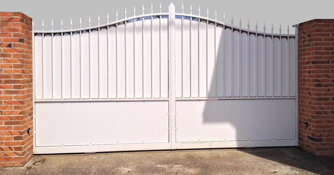 Portail en métal blanc à barreaux verticaux, entre deux piliers en briques.