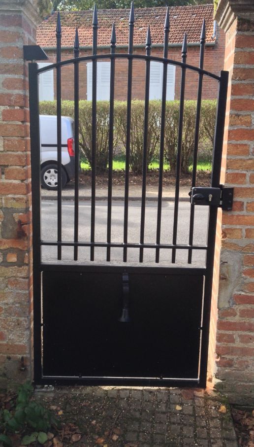 Portail en métal noir dans un mur de briques, avec barreaux verticaux et panneau inférieur plein.