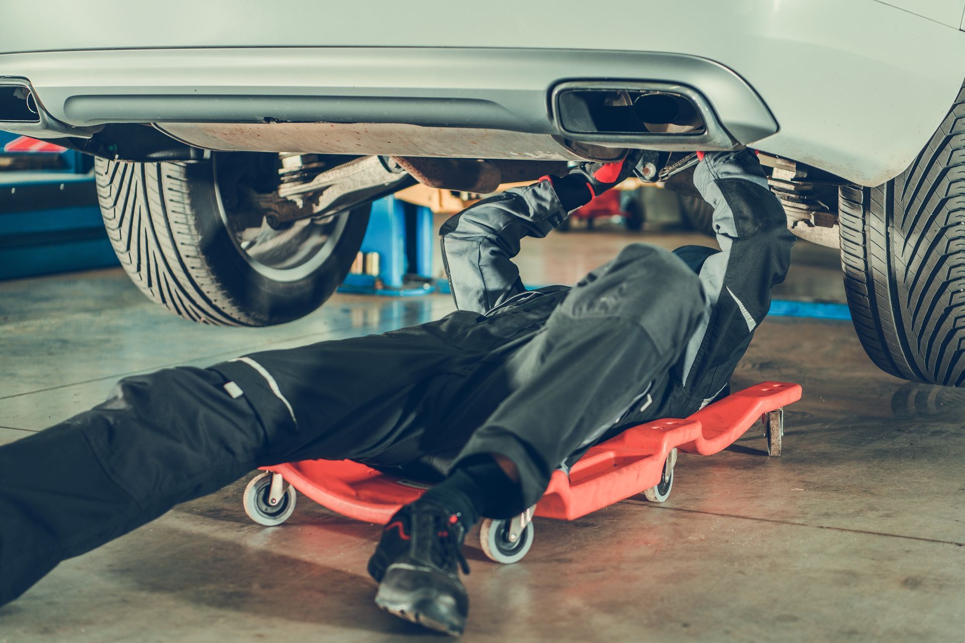 Un mécanicien, installé sur un chariot à roulettes rouge, travaille sous une voiture dans un garage, sur le châssis.