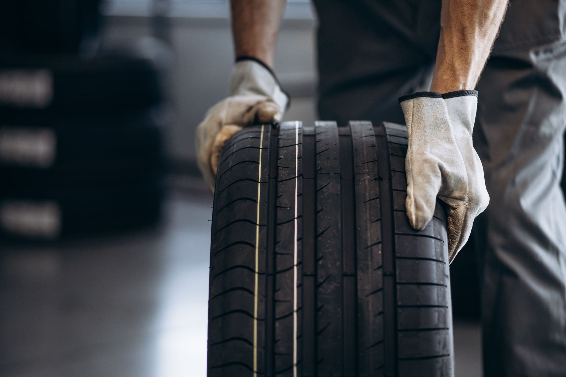 Des mains tiennent deux pneus de voiture dressés dans un garage ou un atelier, avec un véhicule flou en arrière-plan.