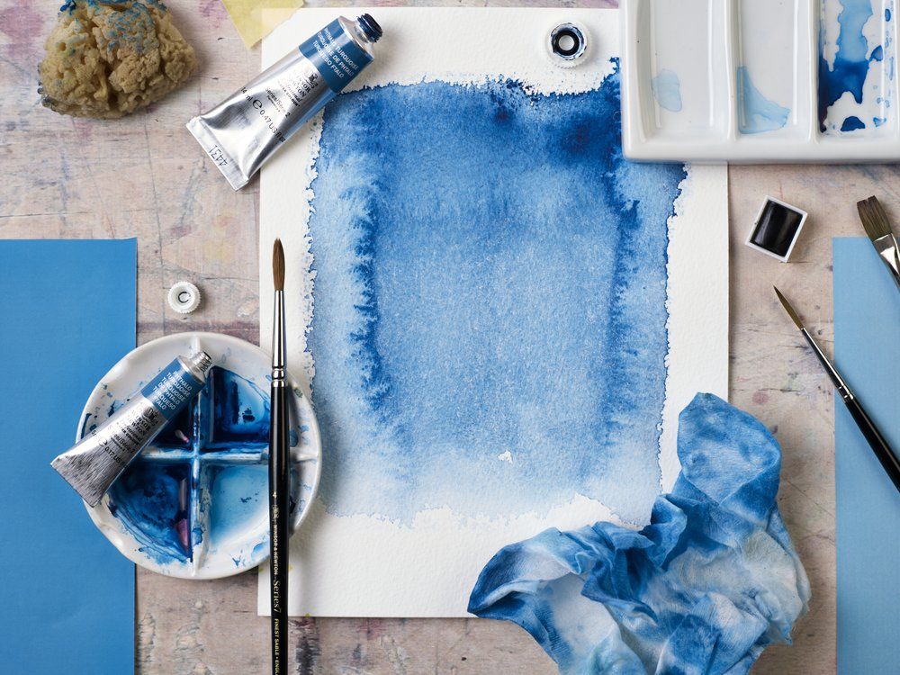 Du matériel d'art, notamment de la peinture aquarelle bleue, un pinceau, une palette et une feuille de papier teintée en bleu, disposés sur un espace de travail.