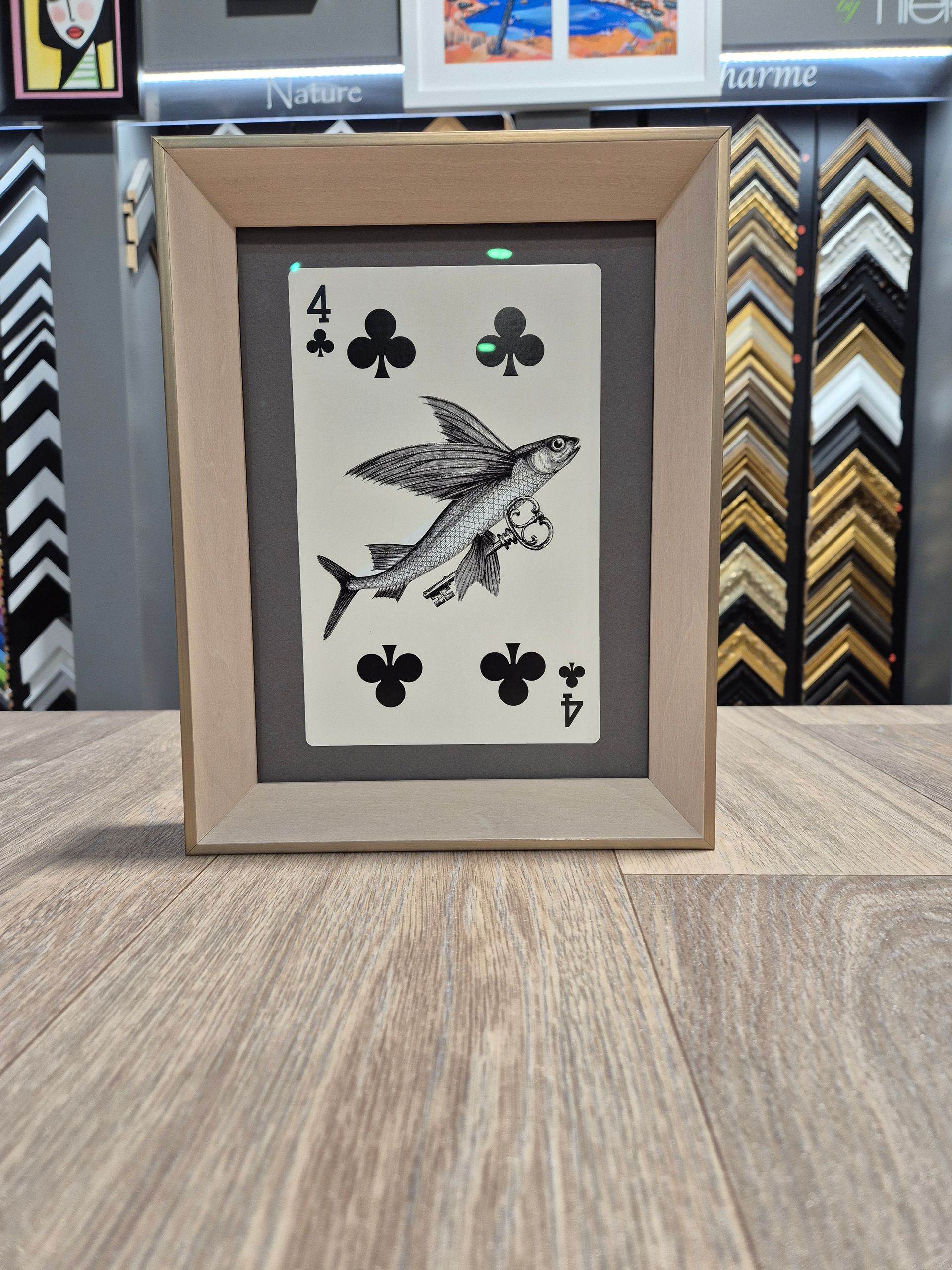 Une carte à jouer 4 de trèfle encadrée, ornée d'une illustration en noir et blanc d'un poisson volant, est présentée sur un comptoir en bois.