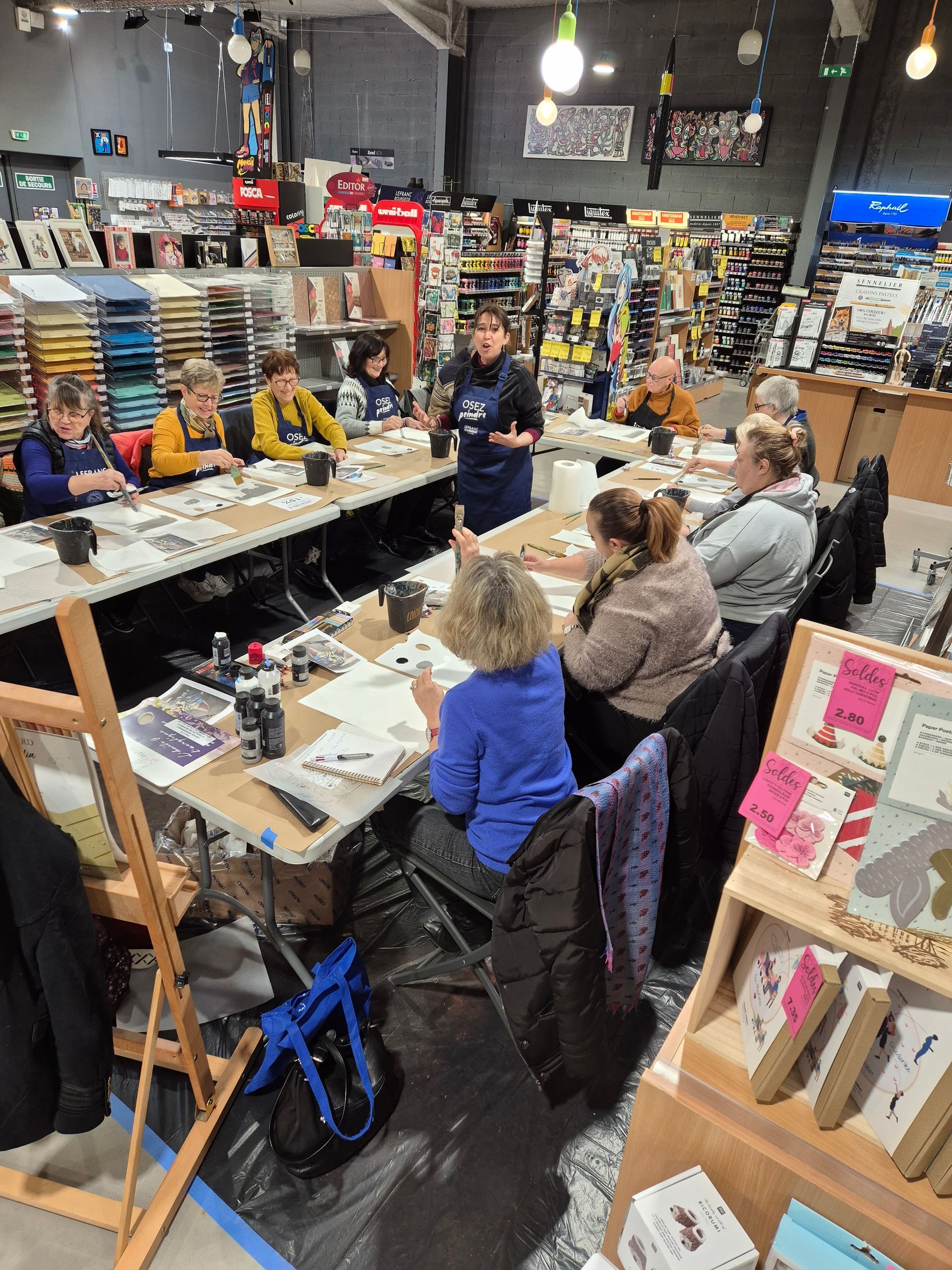 Dans un magasin de fournitures artistiques, un groupe de personnes portant des tabliers est assis à une longue table pour participer à un atelier de peinture.