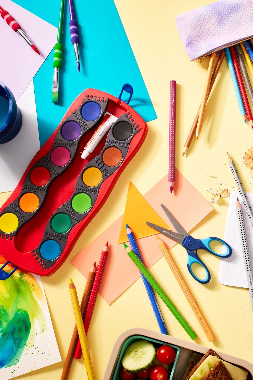 Un espace de travail artistique coloré avec des aquarelles, des crayons de couleur, des ciseaux, du papier et une boîte à lunch contenant des légumes.