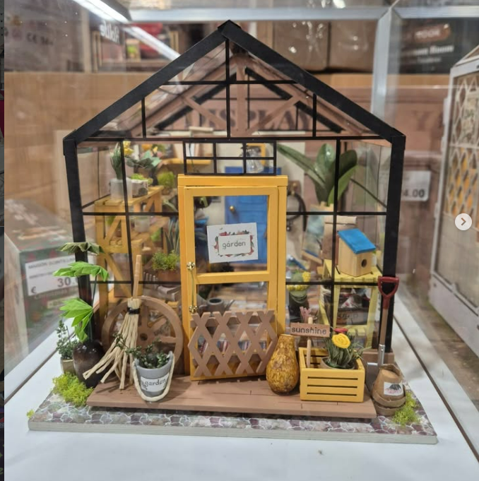 Diorama miniature d'une serre avec porte jaune, plantes et outils de jardinage.