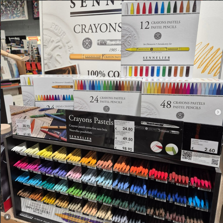 Présentoir de crayons de couleur et de pastels Sennelier dans un magasin. Des boîtes de différentes tailles sont disposées au-dessus de rangées de crayons de couleur.