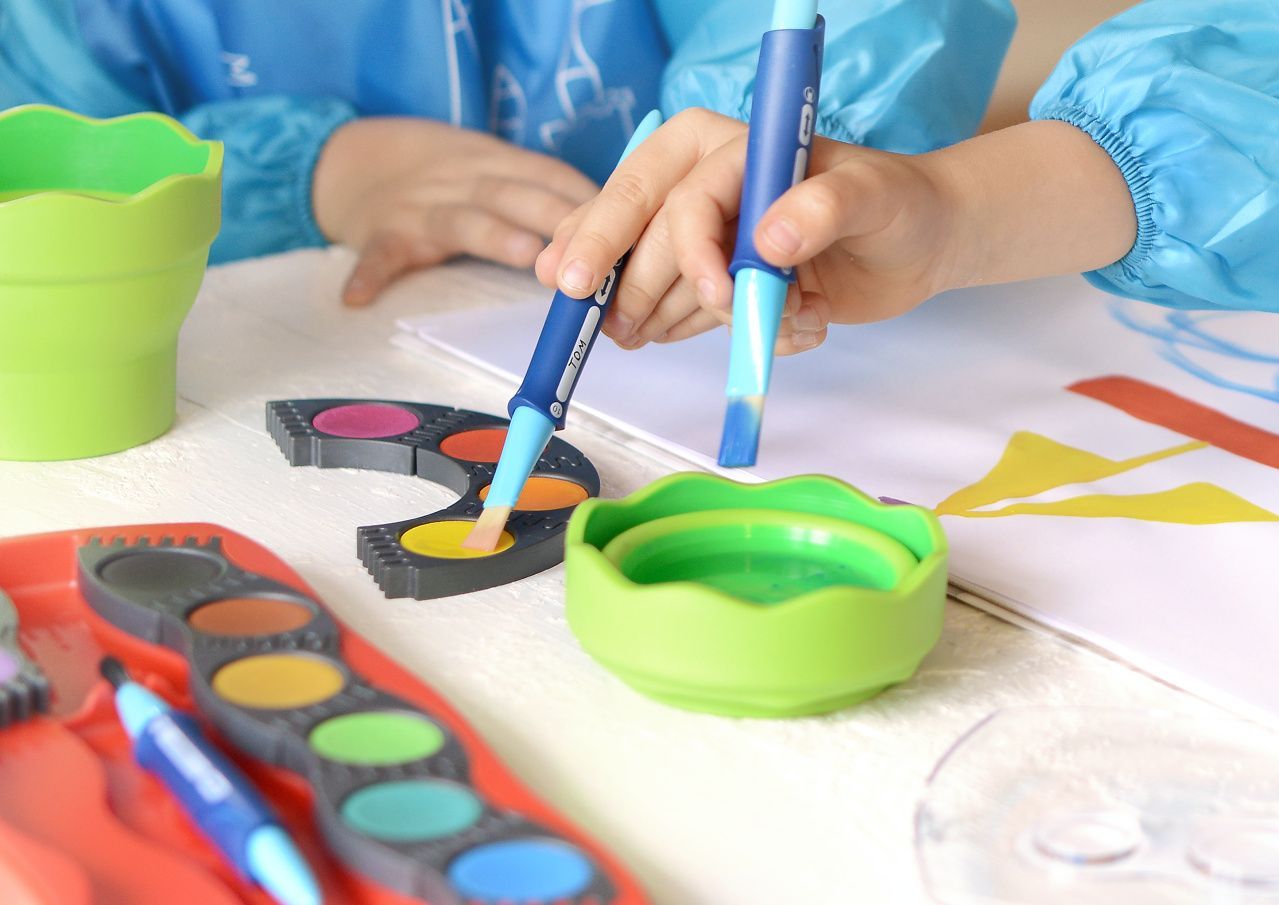 Une personne utilise un pinceau bleu pour prélever de la peinture aquarelle jaune sur une palette en forme de croissant placée à côté d'un gobelet d'eau vert.