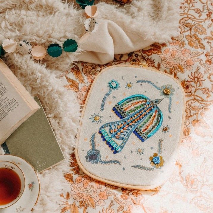 Un tambour à broder à motif d'oiseaux repose sur un textile à motifs floraux, à côté d'une tasse de thé et d'un livre ouvert.