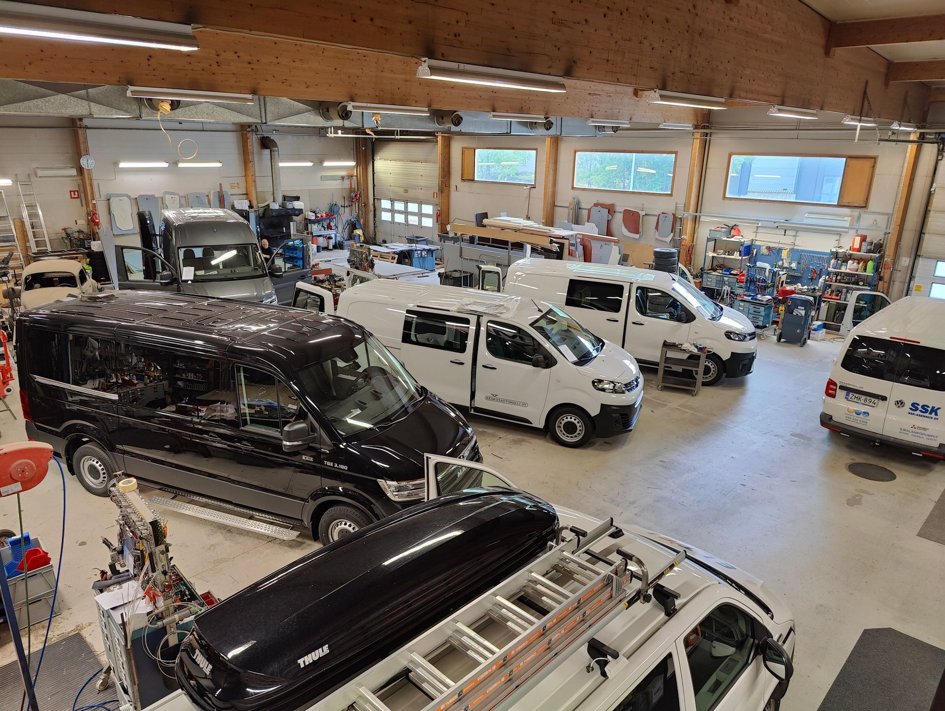 Autokorjaamo, jossa on useita ajoneuvoja, kuten pakettiautoja ja valkoinen katumaasturi, suuren korjaamorakennuksen sisällä