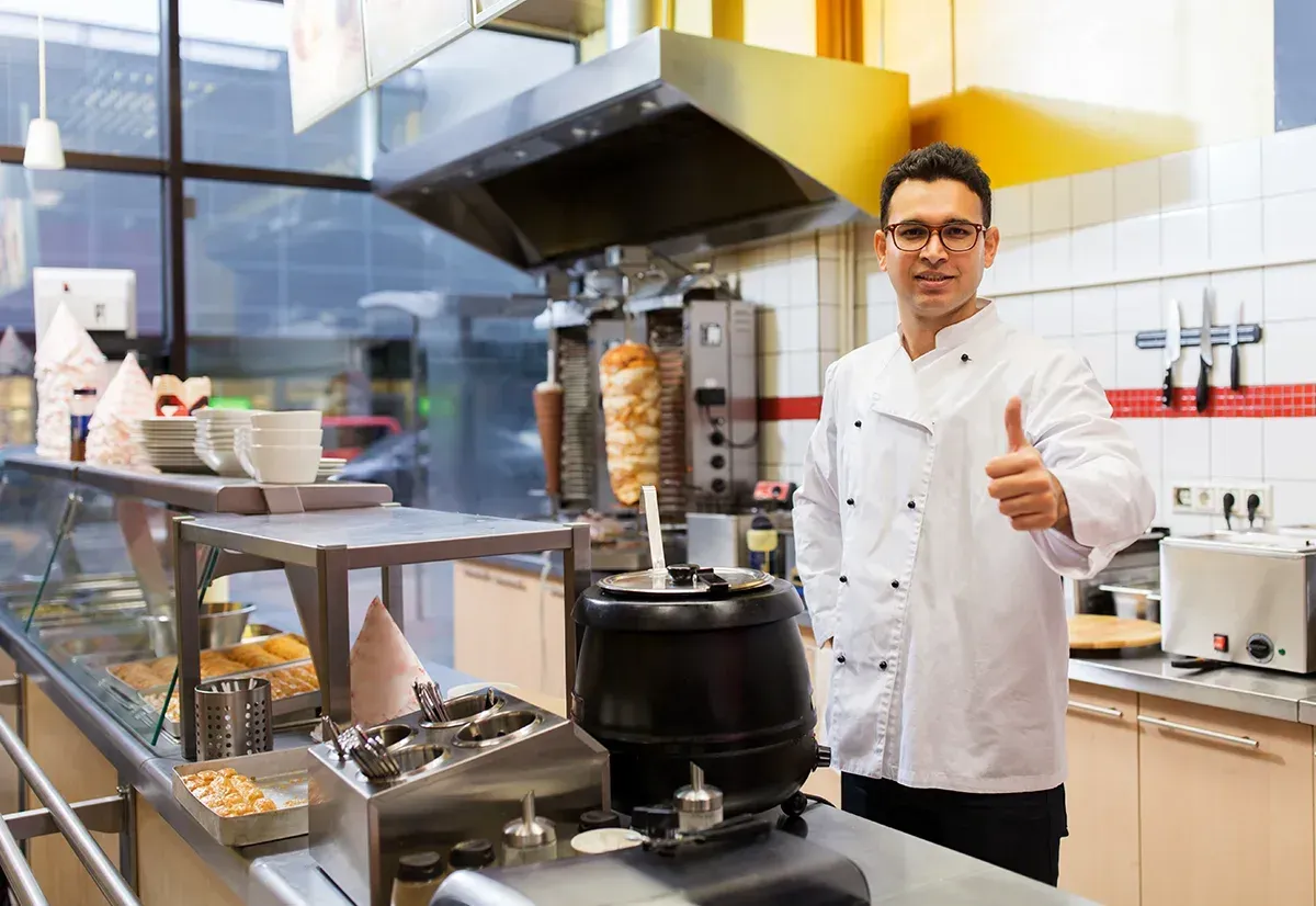 Ein Koch im weißen Kittel zeigt in einer Großküche den Daumen nach oben; im Hintergrund ist ein sich drehender Kebabspieß zu sehen.