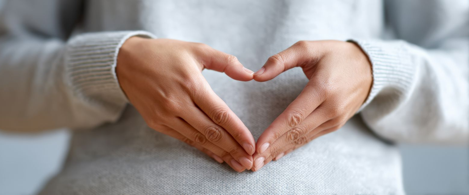 Personne formant un cœur avec ses mains sur son torse, portant un pull gris.
