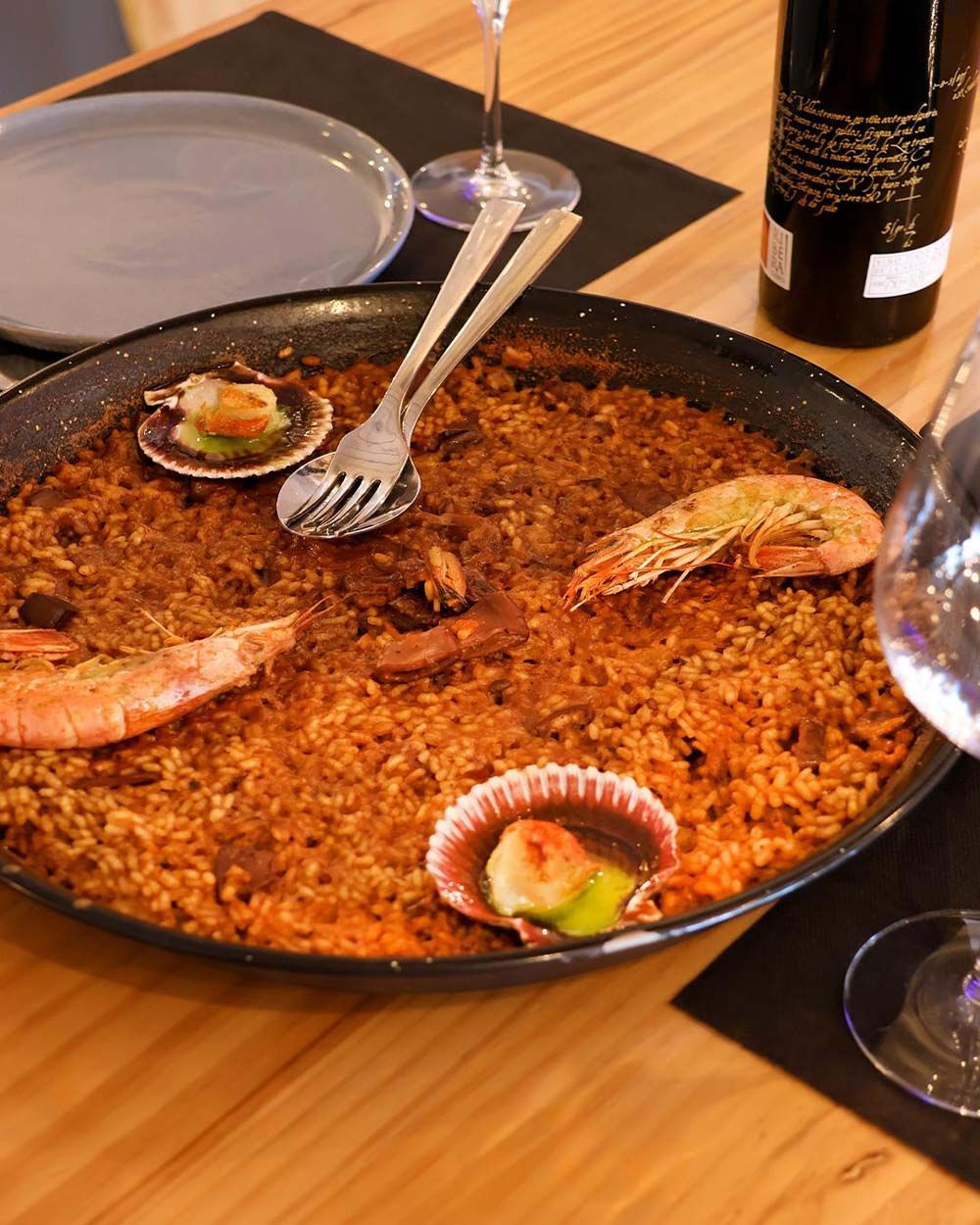 Una sartén con comida y una botella de vino al fondo.
