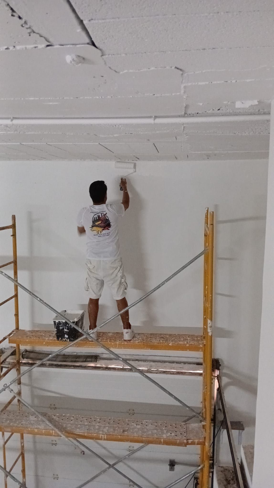 Una persona pinta un techo blanco mientras está de pie sobre un andamio amarillo en una habitación.