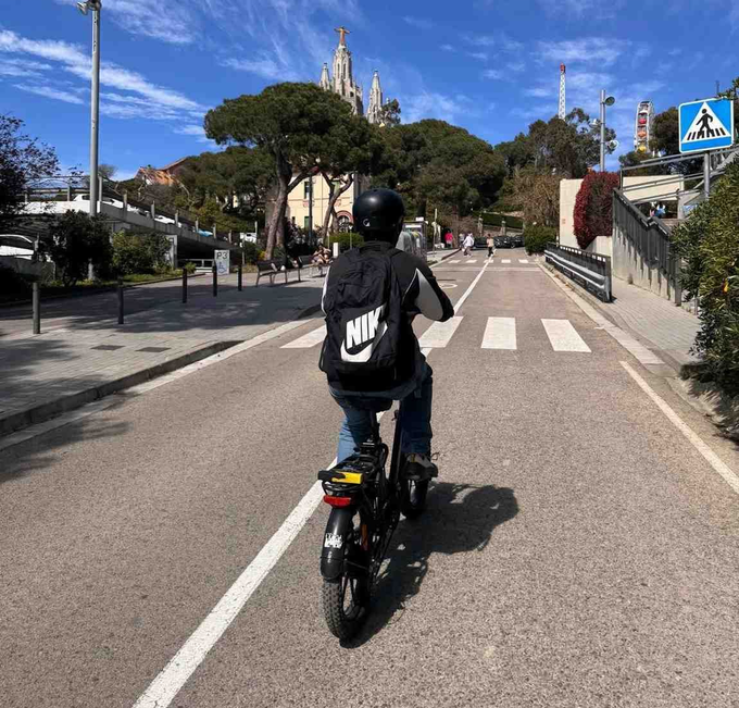 Ciclista subiendo una cuesta en una carretera hacia una iglesia, con un paso de cebra y cielo azul.