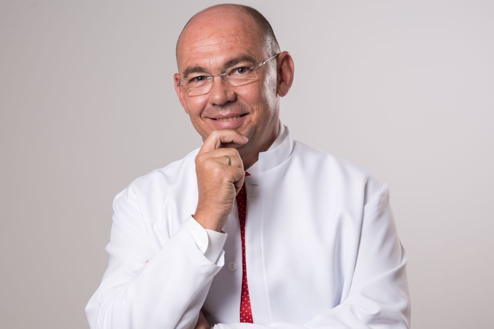 Un hombre con una bata blanca y corbata roja sonríe con la mano en la barbilla.