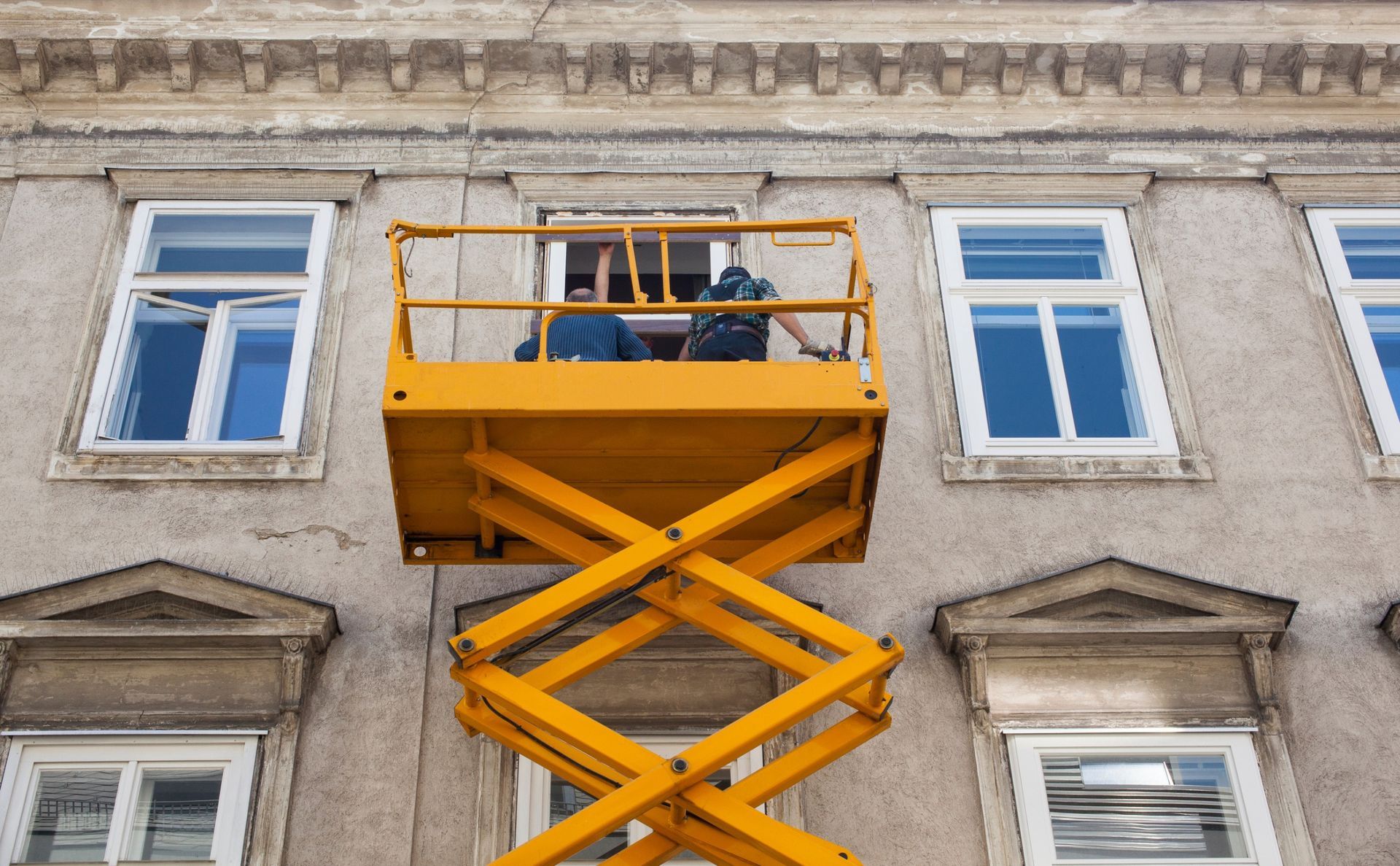 Un hombre está parado en un elevador de tijera amarillo frente a un edificio.