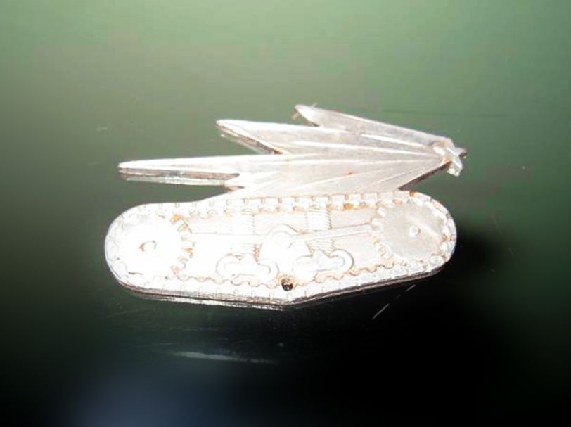 Une broche métallique argentée en forme de char d'assaut ailé, se détachant sur une surface vert foncé réfléchissante.