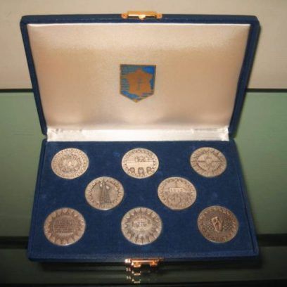 Un coffret de présentation en velours bleu contenant huit médaillons circulaires en argent, avec un petit blason décoratif sur le couvercle.