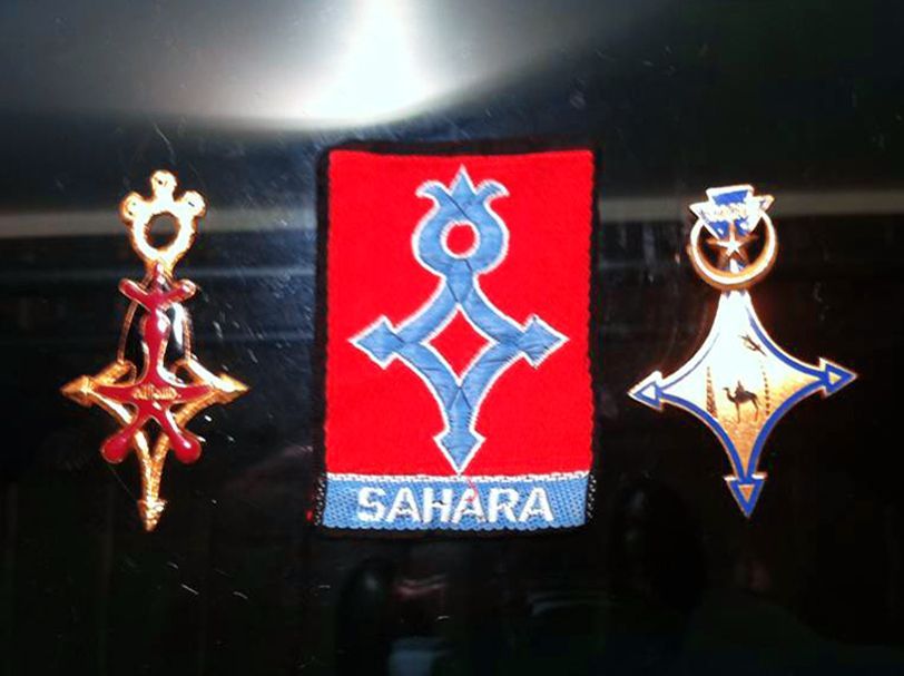 Un écusson central rouge orné d'une croix touareg bleue et de l'inscription SAHARA, flanqué de deux pendentifs métalliques assortis en forme de croix touareg.