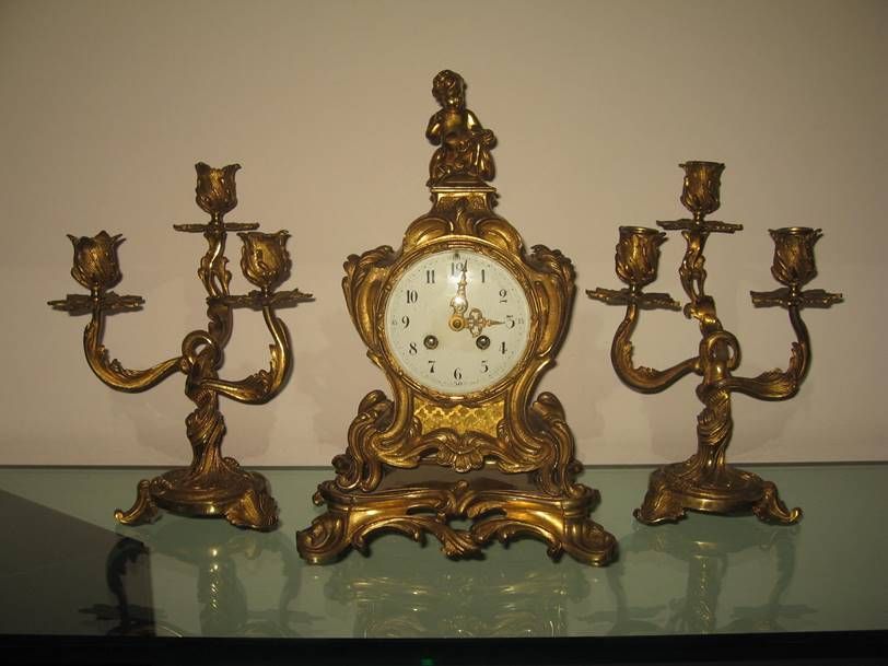 Une pendule de cheminée française ornée, de couleur dorée, flanquée de deux candélabres à trois bougies assortis, posée sur un plateau en verre.