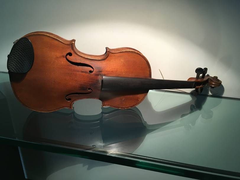 Un violon en bois repose sur une étagère en verre, devant un mur blanc éclairé d'une lumière douce et chaleureuse.